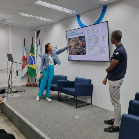 A imagem em destaque mostra a residente Andressa Santos mostrando o mapa da sua unidade na tela de uma tv ao centro. Ao seu lado de costas está seu colega de unidade assistindo a sua apresentação. 