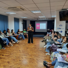 Foto de uma sala de aula. Os alunos estão sentados em círculo e a professora está ao centro da imagem. 